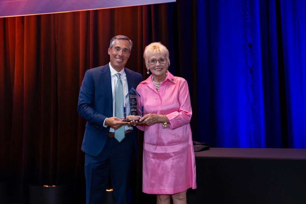 Marilyn Hatley, the former Mayor of North Myrtle Beach, smiles for award with Todd Setzer from A&I Fire and Water Restoration. Marilyn is wearing a pink dress, Todd is dressed in a blue suit and tie.